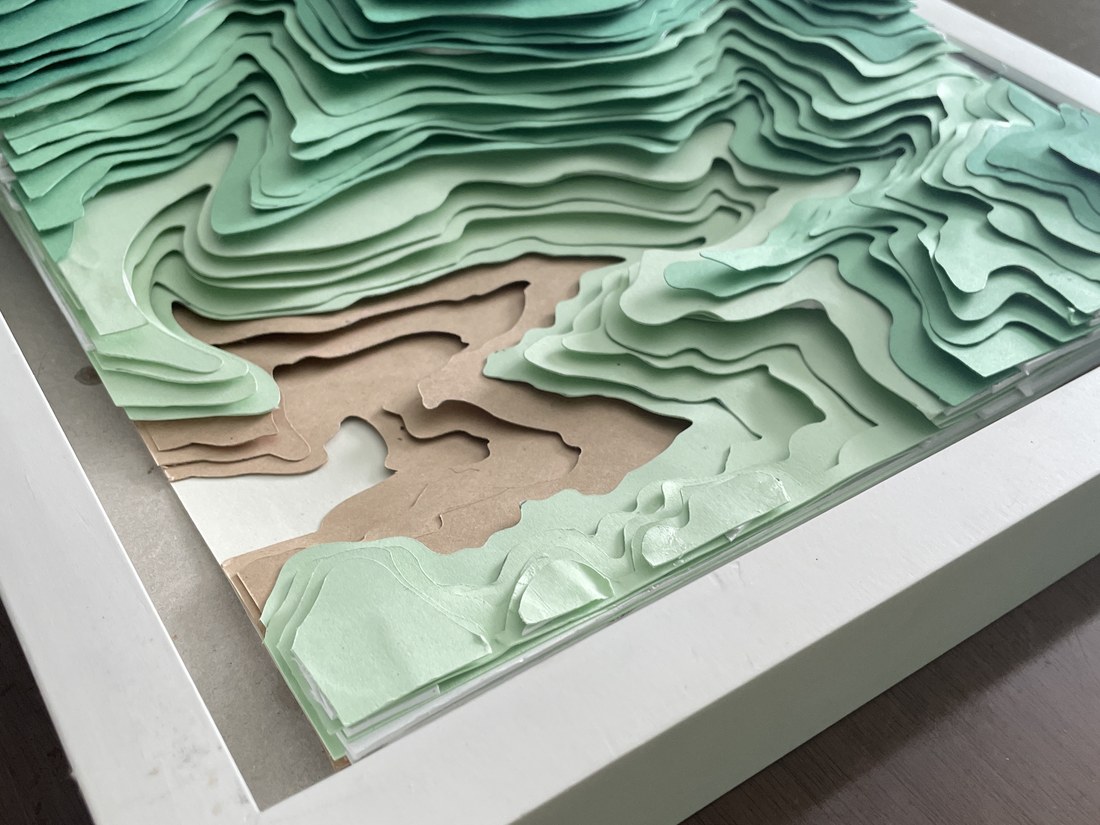 A low-angle view of the framed shadowbox, the layered cardstock terrain casting real shadow inside the white frame